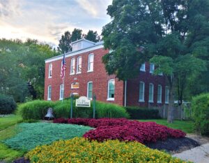 Two story brick building with twelve windows cased in white trim with a variety of green trees and bushes around.